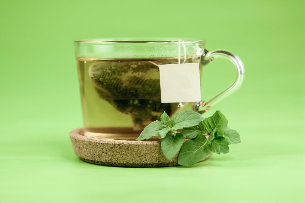 A cup of spearmint tea with a teabag and some spearmint leaves by the side.